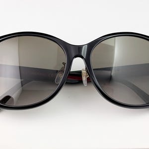 May include: Black oversized sunglasses with dark lenses. The sunglasses have a thick, rounded frame and the brand name "GUCCI" is visible on the left lens. The temples are black with green tips.