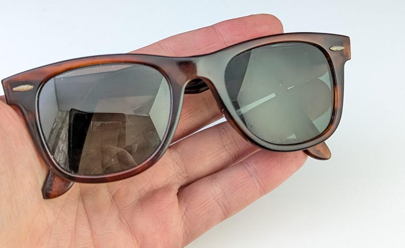 May include: Brown tortoiseshell sunglasses with dark lenses. The sunglasses have a classic, rectangular frame design with a keyhole bridge. The lenses are reflective, and the arms have a slight curve for a comfortable fit.