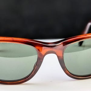May include: Brown tortoiseshell sunglasses with green lenses. The classic frame features a rounded shape and a keyhole bridge. The temples and frame have a mottled pattern. The sunglasses are set against a white surface and a dark background.