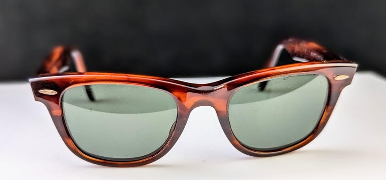 May include: Brown tortoiseshell sunglasses with green lenses. The classic frame features a rounded shape and a keyhole bridge. The temples and frame have a mottled pattern. The sunglasses are set against a white surface and a dark background.