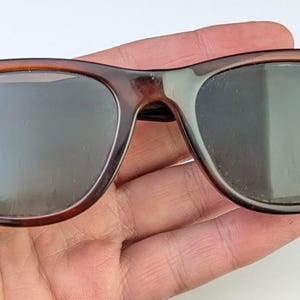 May include: Brown tortoiseshell sunglasses with dark lenses. The classic frame has a rectangular shape with rounded edges. The sunglasses are held in a hand, showcasing the details of the frame and lenses.