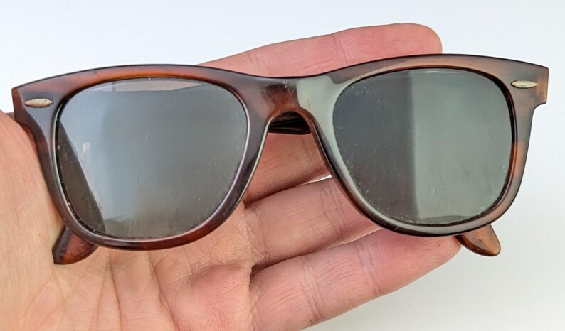 May include: Brown tortoiseshell sunglasses with dark lenses. The classic frame has a rectangular shape with rounded edges. The sunglasses are held in a hand, showcasing the details of the frame and lenses.