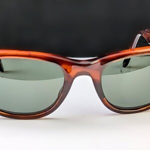 May include: Brown tortoiseshell sunglasses with green lenses. The sunglasses have a classic, retro design with a thick frame and a keyhole bridge. The temples have a textured detail.