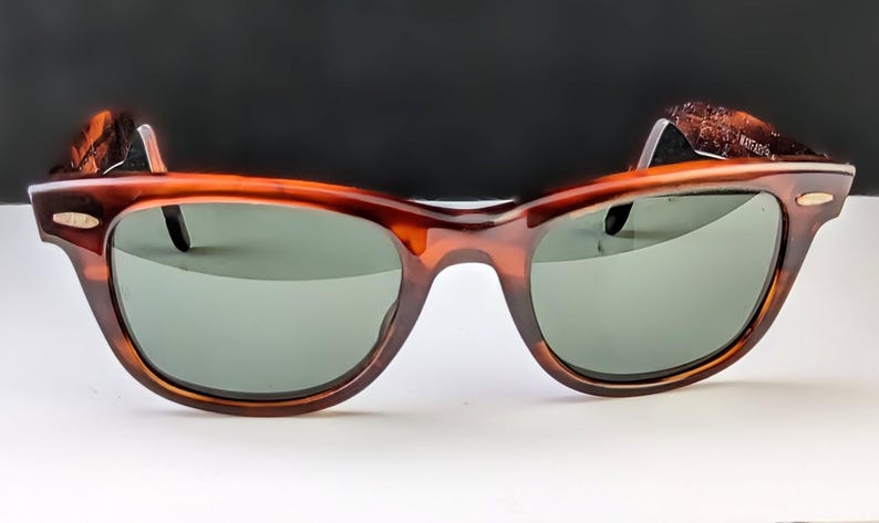 May include: Brown tortoiseshell sunglasses with green lenses. The sunglasses have a classic, retro design with a thick frame and a keyhole bridge. The temples have a textured detail.