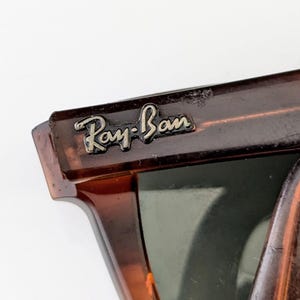 May include: Close-up of Ray-Ban sunglasses. The frame has a tortoiseshell pattern, with a dark brown and amber colour scheme. The Ray-Ban logo is visible on the upper left corner of the frame in silver lettering. The lenses are a dark green colour.