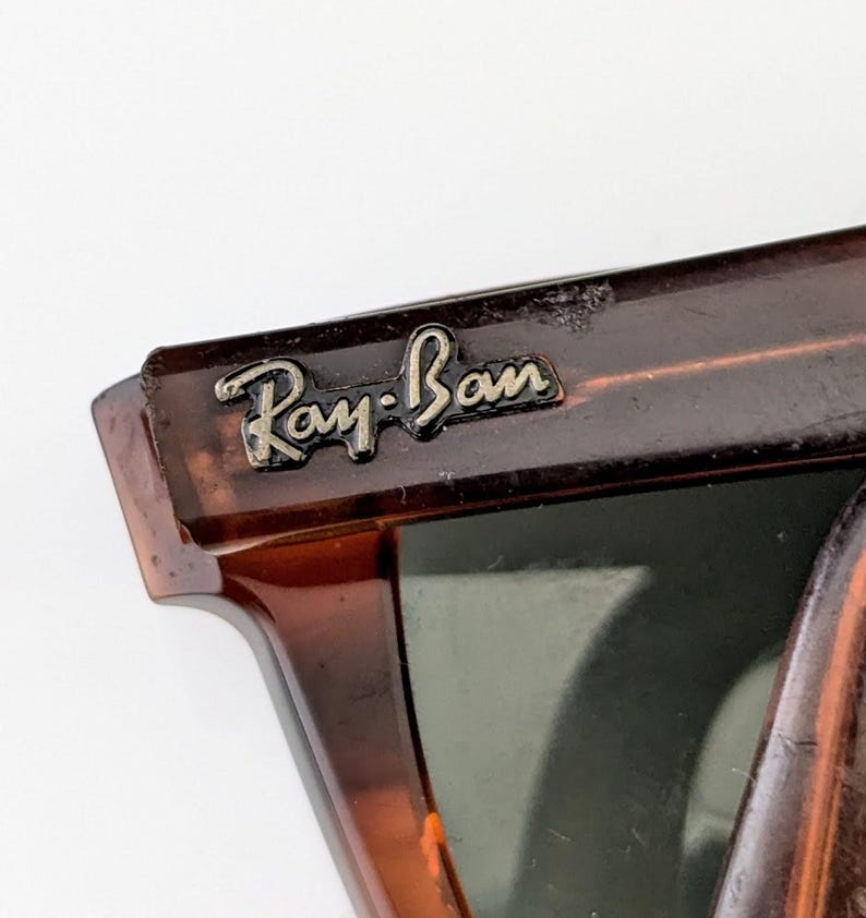 May include: Close-up of Ray-Ban sunglasses. The frame has a tortoiseshell pattern, with a dark brown and amber colour scheme. The Ray-Ban logo is visible on the upper left corner of the frame in silver lettering. The lenses are a dark green colour.