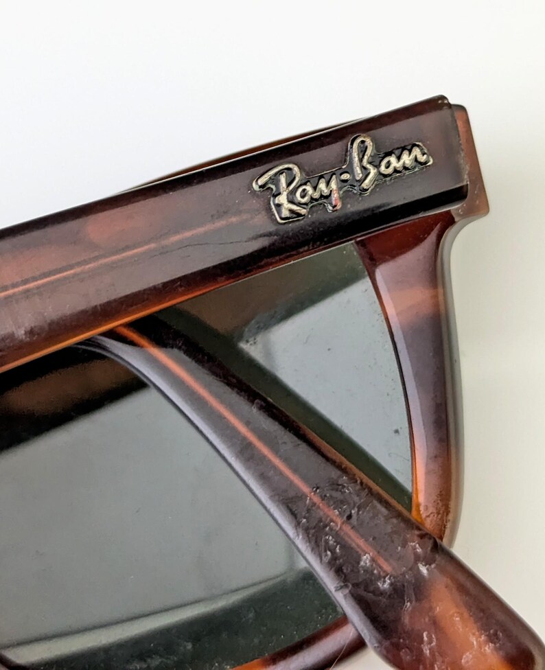 May include: Close-up of Ray-Ban sunglasses with a tortoiseshell frame. The sunglasses have dark lenses and the Ray-Ban logo is visible on the top corner of the frame. The frame is a mix of brown and amber colours.