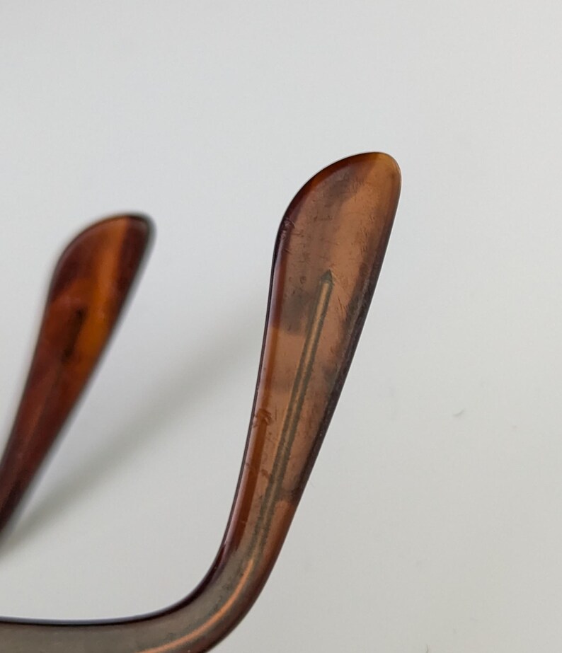 May include: Close-up of the temple arms of eyeglasses. The arms are a translucent brown with a marbled effect. The shape is curved, and the tips are rounded. The background is a plain white.