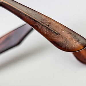 May include: Close-up of the arm of a pair of tortoiseshell spectacles. The arm is a warm brown colour with a mottled pattern. The end of the arm curves downward, and the hinge is visible. The spectacles are set against a white background.