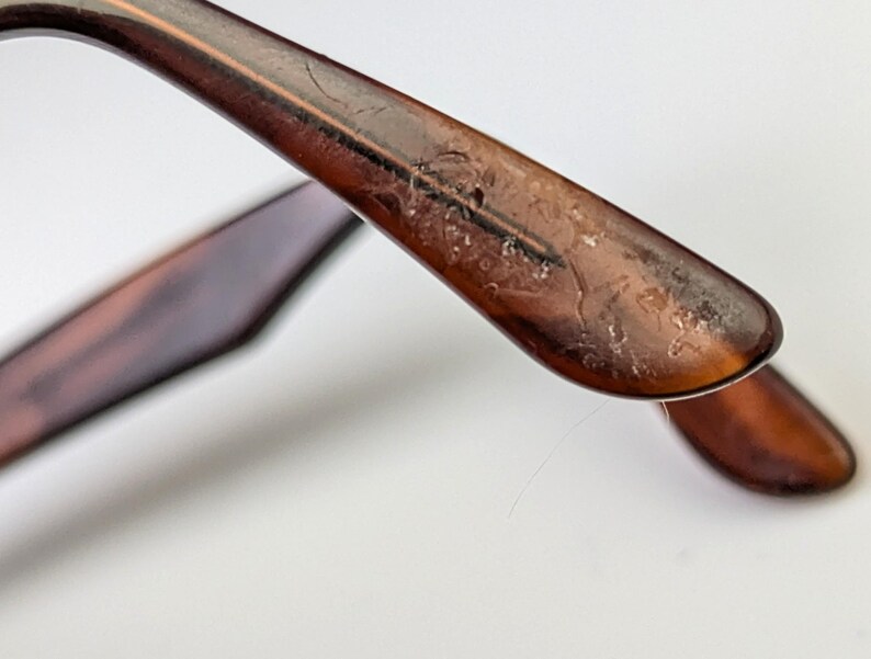 May include: Close-up of the arm of a pair of tortoiseshell spectacles. The arm is a warm brown colour with a mottled pattern. The end of the arm curves downward, and the hinge is visible. The spectacles are set against a white background.