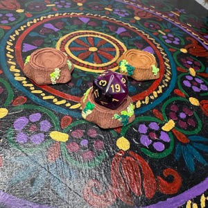 May include: A purple and gold D20 die rests on a brown, tree stump-shaped dice holder, adorned with green and yellow accents. Two additional holders are visible. The items are set on a black surface with a colorful, circular floral design.