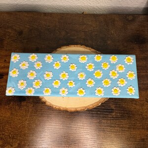 May include: A rectangular canvas painting with a light blue background and a pattern of white daisies with yellow centers.