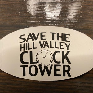 May include: Oval sticker with the text "SAVE THE HILL VALLEY CLOCK TOWER" in black, with a clock graphic. The sticker is white with a glossy finish.