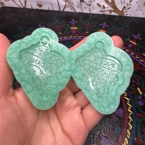 May include: Two light green resin molds in a hand. Each mold is shaped like a decorative tag with the text "ASK GUIDANCE" and "FOR MAGIC" in white. The molds have a glittery finish and are designed for crafting.