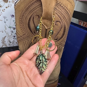 May include: A tan leather cowboy boot with decorative stitching. A gold chain with a leaf charm and beads hangs from the boot. The leaf charm has a green and purple iridescent finish.