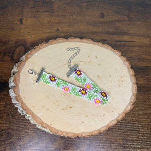 May include: A white beaded bracelet with a floral pattern in pink, green, and yellow. The bracelet has silver clasps.