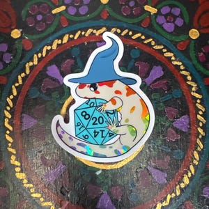 May include: A sticker featuring a cartoon lizard wearing a blue wizard hat and holding a holographic d20 die. The lizard has orange and white spots. The background is a colorful, ornate design.