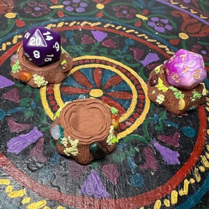 May include: Three miniature, handcrafted dice holders, each resembling a tree stump, are displayed. Two holders feature a purple and pink d20 die, while the third is empty. The holders are adorned with small, colorful details, set against a decorative, patterned background.