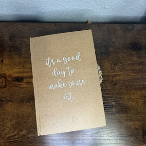 May include: A brown notebook with the text "it's a good day to make some art." written in white on the front cover.
