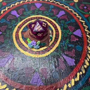 May include: A purple D20 die rests on a small, decorated base with blue and purple flowers. The die displays white numbers against a translucent purple background. The base sits on a dark, ornate surface with floral and geometric patterns.