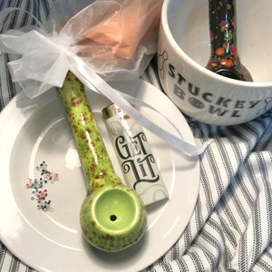 Puede incluir: Una pipa de cerámica verde y marrón moteada con un cuenco redondo, un encendedor blanco con el texto "GET LIT" y un cuenco blanco con el texto "STUCKEY BOWL". Los artículos están colocados sobre un plato blanco y una tela a rayas.