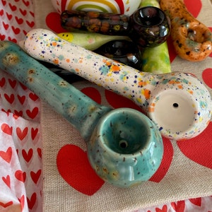 May include: A collection of ceramic pipes in various colors and designs. One is teal with speckled accents, another is white with colorful splatters, and others feature brown, orange, and green hues. The pipes are arranged on a red heart-patterned surface.