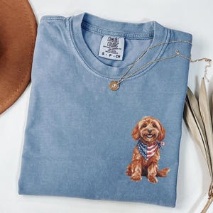 May include: A light blue Comfort Colors t-shirt with a graphic of a brown dog wearing an American flag bandana. A gold necklace and a brown hat are also in the image. The shirt is size S-P-CH.
