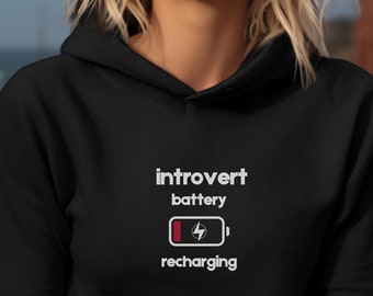 Puede incluir: Sudadera con capucha negra con texto blanco que dice "introvert battery recharging" y un icono de batería blanco con una barra roja.