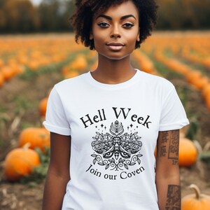 Op de afbeelding: Wit T-shirt met een zwart grafisch ontwerp met een nachtvlinder met vleugels en de tekst "Hell Week Join our Coven".