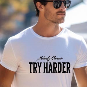 May include: White crew neck t-shirt with the text "Nobody Cares TRY HARDER" in black. The shirt is worn with gray pants. The person is wearing sunglasses and a watch.