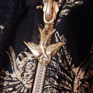 May include: Close-up of a gold zipper pull in the shape of a bird, attached to a black fabric with a gold floral pattern. The zipper is partially open, revealing the silver teeth.
