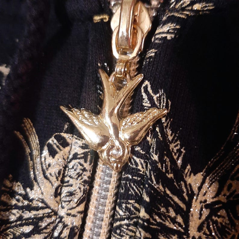 May include: Close-up of a gold zipper pull in the shape of a bird, attached to a black fabric with a gold floral pattern. The zipper is partially open, revealing the silver teeth.