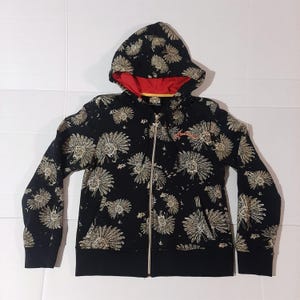 May include: Black zip-up hoodie with a floral pattern. The hoodie features a red lining in the hood and a gold zipper. The floral design is in a light tan color. The cuffs and bottom hem are solid black.