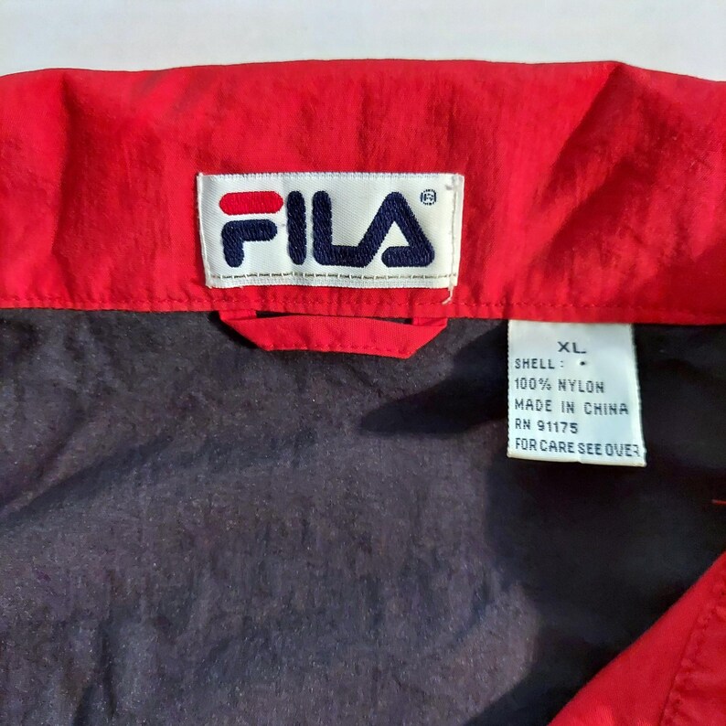 Pu&ograve; includere: Primo piano di una giacca Fila rossa e blu navy. Il logo Fila &egrave; visualizzato su un'etichetta bianca. La giacca &egrave; XL e realizzata in 100% nylon. L'etichetta include anche il testo "MADE IN CHINA" e "FOR CARE SEE OVER."