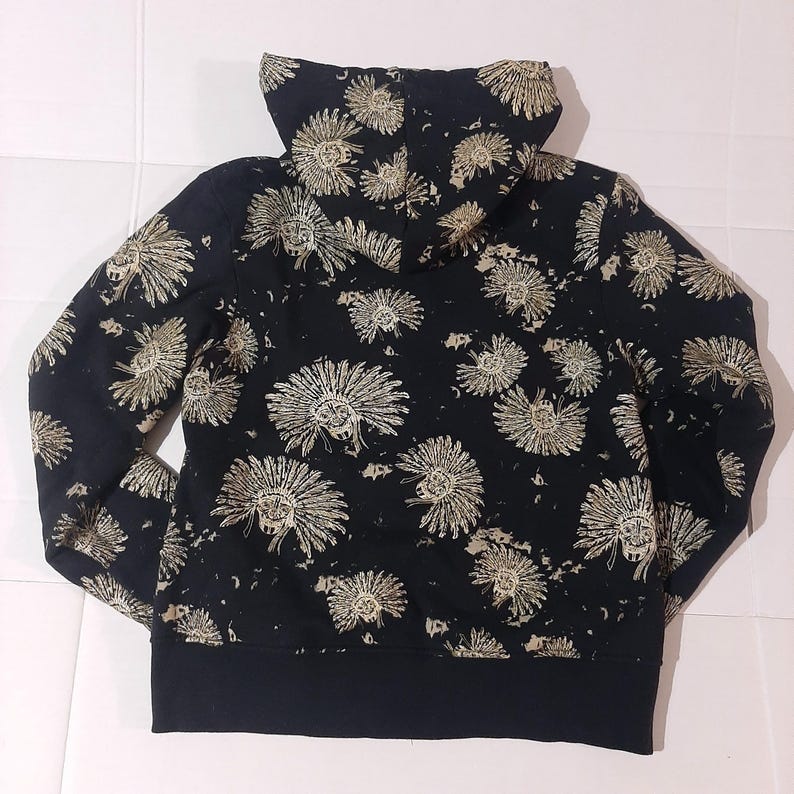 May include: Black hoodie with a repeating pattern of tan Native American headdresses. The hoodie has a black ribbed hem and cuffs, and a lined hood. The design is printed on a black background.