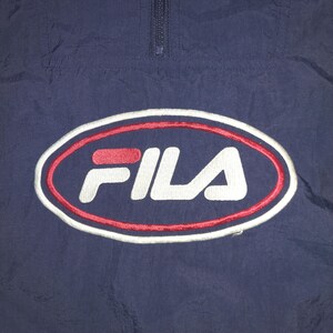 Pu&ograve; includere: Giacca sportiva blu navy con logo FILA. Il logo &egrave; un design ovale con una combinazione di colori rosso e bianco. La giacca ha una chiusura con zip a un quarto sul collo.