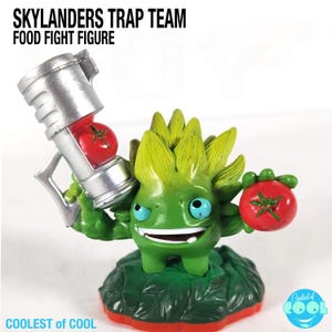 May include: A green Skylanders Trap Team Food Fight figure with a lime green spiky head and blue eyes. The figure holds a silver weapon with a red tomato inside and a red tomato in its other hand. The figure is on a green leafy base.