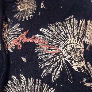 May include: Close-up of a navy blue fabric with a repeating pattern of skull headdresses and floral designs in off-white. The word "Arise" is embroidered in red script. The overall design has a vintage, bohemian aesthetic.