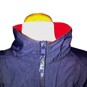 Pu&ograve; includere: Primo piano di una giacca blu navy con fodera del colletto rossa. La giacca ha un collo alto e una cerniera completa. Un'etichetta blu e bianca con il nome del marchio &egrave; attaccata alla cerniera.