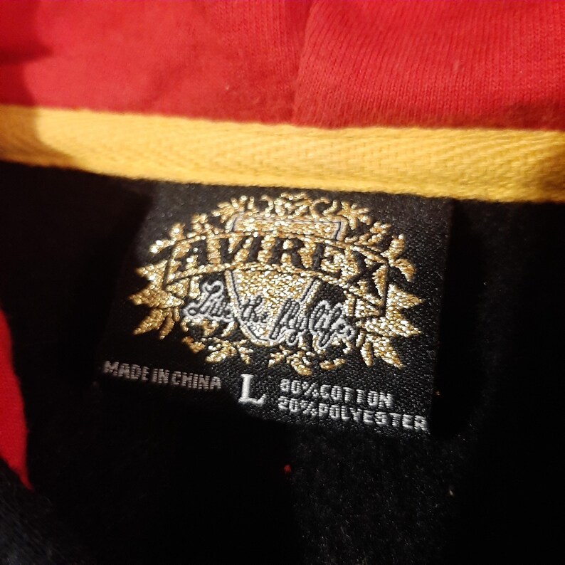 May include: Close-up of a black label with gold lettering and the brand name "AVIREX". The label also includes the text "Live the Fly Life", "Made in China", and the size "L". The label is on a black garment with red and yellow accents.