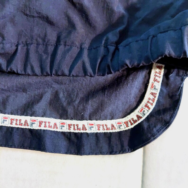 Pu&ograve; includere: Primo piano di un capo sportivo blu navy. Il capo presenta un bordo bianco e rosso con il nome del marchio "FILA" ripetuto. Il tessuto sembra essere un materiale leggero, forse nylon.