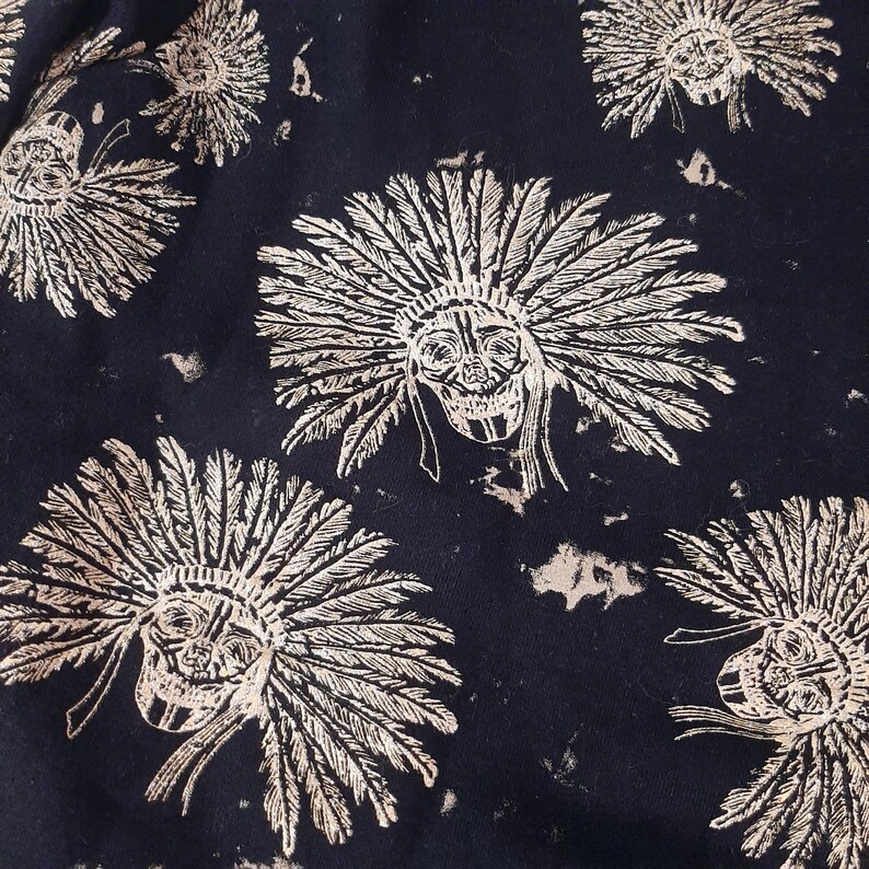 May include: Dark navy fabric with a repeating pattern of skull faces wearing feathered headdresses. The skulls are outlined in a light beige color, with detailed feather designs. The overall design has a distressed, vintage look.