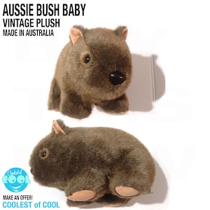 May include: A brown plush toy resembling a wombat with dark eyes, a black nose, and light tan inner ears. The text AUSSIE BUSH BABY VINTAGE PLUSH MADE IN AUSTRALIA appears at the top, and a circular logo with the text COOLEST OF COOL is visible.
