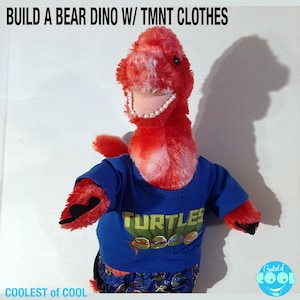 May include: A red and white plush dinosaur wearing a blue t-shirt with the Teenage Mutant Ninja Turtles logo and matching pajama pants. The text "TURTLES" is printed on the shirt. The dinosaur is smiling and has a shadow behind it.