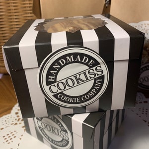 May include: Two black and white striped boxes with a clear window on top, stacked on top of each other. Each box has a round sticker that says "HANDMADE COOKISS COOKIE COMPANY". The boxes are on a white surface.