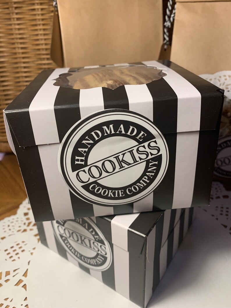 May include: Two black and white striped boxes with a clear window on top, stacked on top of each other. Each box has a round sticker that says "HANDMADE COOKISS COOKIE COMPANY". The boxes are on a white surface.