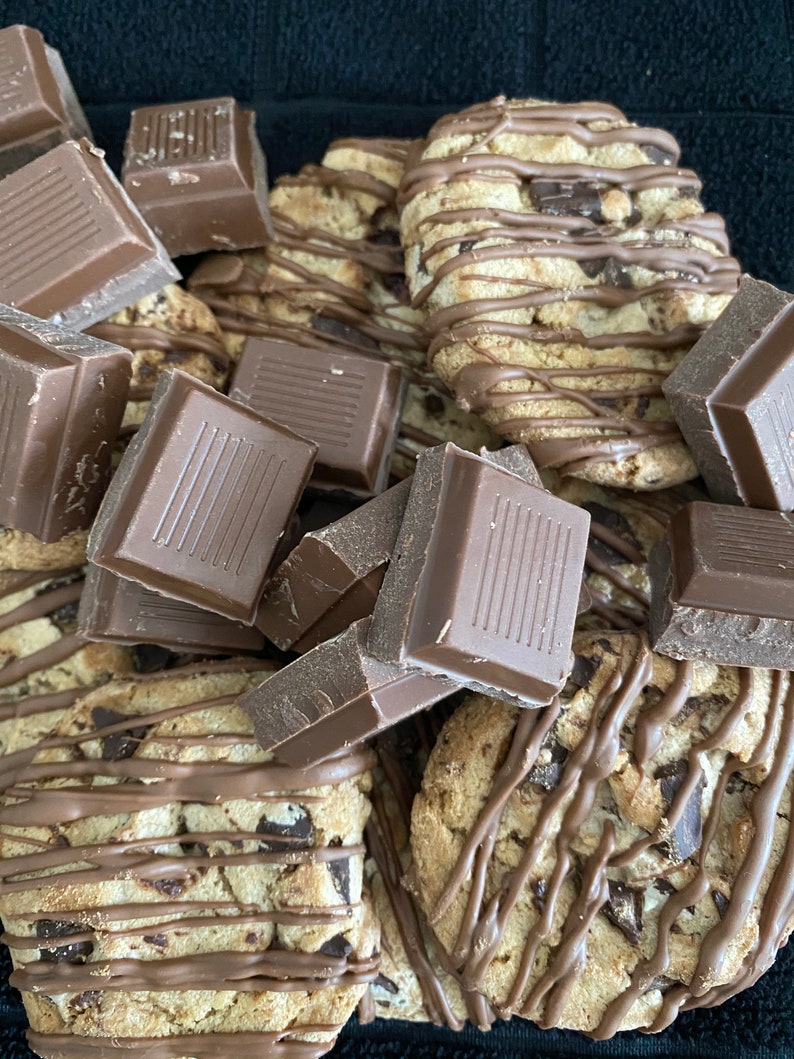 May include: Close-up of chocolate chip cookies drizzled with chocolate and surrounded by chunks of chocolate bars. The cookies have a golden-brown color with visible chocolate chips.