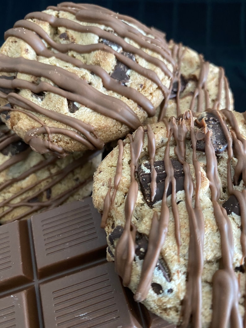 May include: Close-up of several chocolate chip cookies drizzled with chocolate. The cookies have large chocolate chunks and are stacked near a dark chocolate bar. The cookies are a light brown color and the chocolate drizzle is a darker brown.