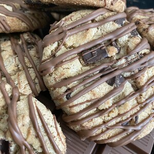 May include: Close-up of several chocolate chip cookies drizzled with chocolate. The cookies are light brown with visible chocolate chips and a textured surface. A few pieces of a chocolate bar are visible in the background.