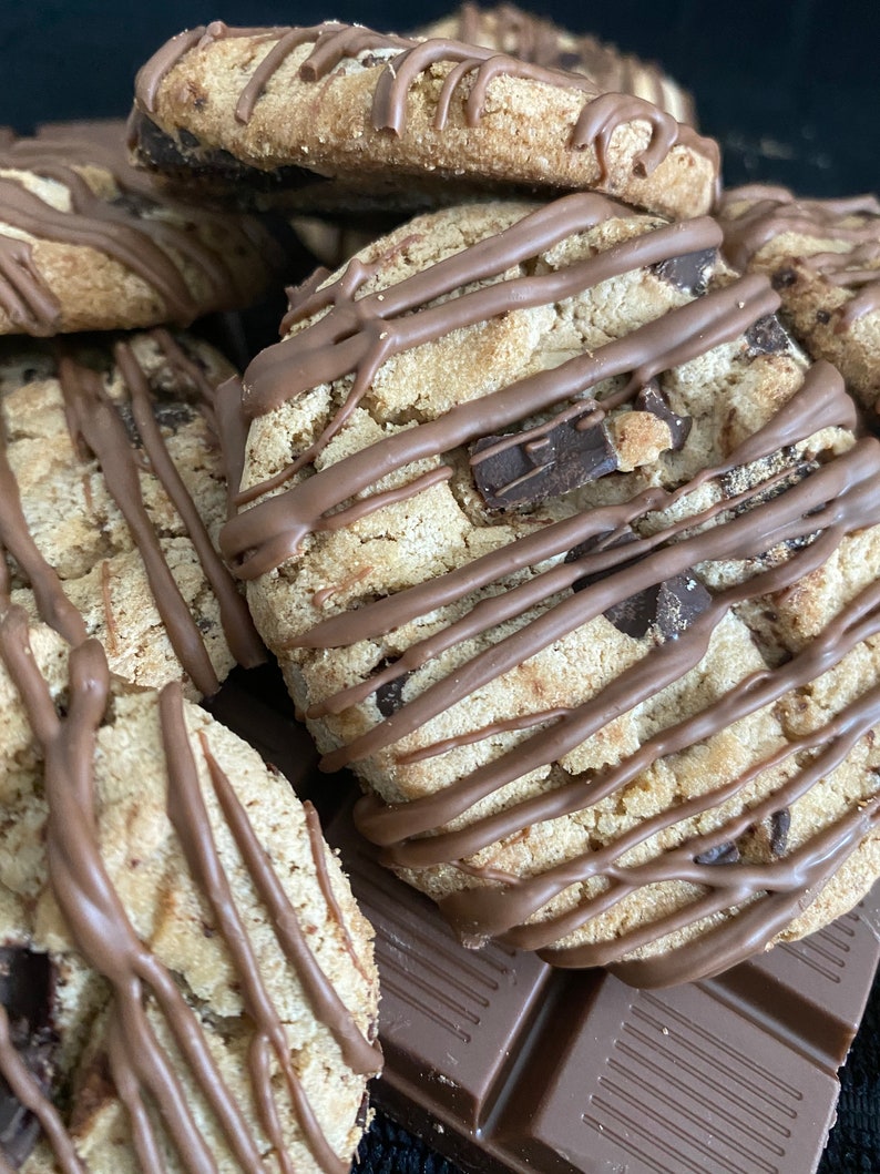 May include: Close-up of several chocolate chip cookies drizzled with chocolate. The cookies are light brown with visible chocolate chips and a textured surface. A few pieces of a chocolate bar are visible in the background.